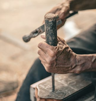 Weathered hands working a hammer and chisel against a slab of stone.