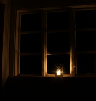 A single lit candle glowing at the bottom of a dark window at night.