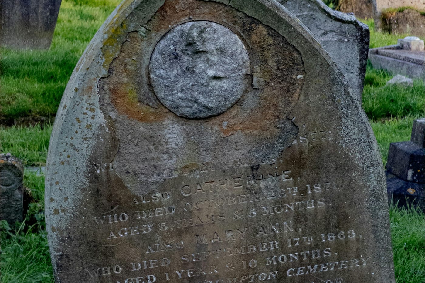 A weathered gravestone with a round medallion above a worn inscription for two young children, standing in green grass.