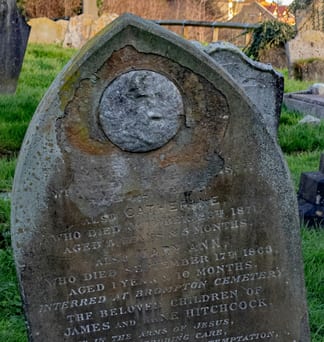 A weathered gravestone with a round medallion above a worn inscription for two young children, standing in green grass.